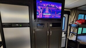 Ein kompakter Wohnraum mit einem Fernseher über einem dunkelgrauen Schrank, einem Etagenbett mit roter Decke und einer kleinen Bedienkonsole unter dem Fernseher.