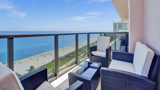 Een balkon met rieten meubels en een glazen tafel, met uitzicht op een strand.