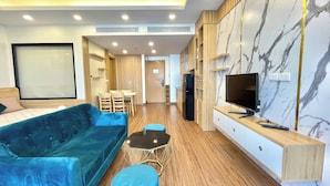 A modern living room with a blue sofa, a small round ottoman, a glass coffee table, a TV on a wooden stand, and a marble-patterned wall. - Dan - FLC Sea Tower Quy Nhon (Quy Nhon Nam)