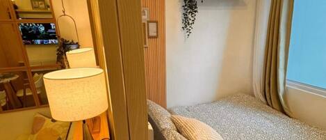 A compact hotel room with a bed, a bedside lamp, a small table, and a mirror.