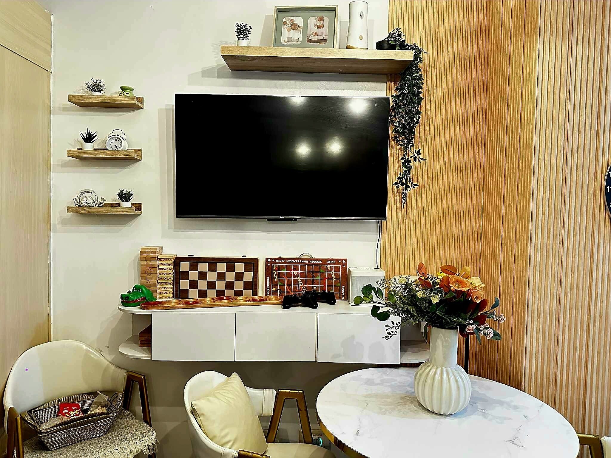 A modern living room with a flat-screen TV, wooden shelves, a chess set, a gaming console, and a dining area with a white table and chairs.