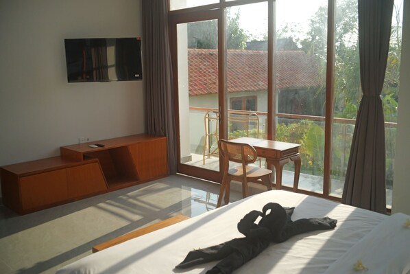 A bedroom with a large bed, a wooden desk, a TV mounted on the wall, and a balcony with a view of the outside. - Ulu Penthouse (Pecatu)