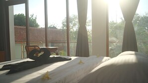 Una stanza con un letto, una sedia e un tavolo. Sono presenti tende e una finestra che si affaccia su alberi e un edificio.