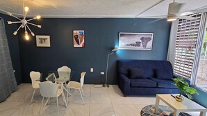 A modern living room with a blue sofa, a glass dining table, and white chairs. There are two framed pictures on the wall, a ceiling light fixture, and a window with blinds. - The Central Comfort in Kingston (Kingston)