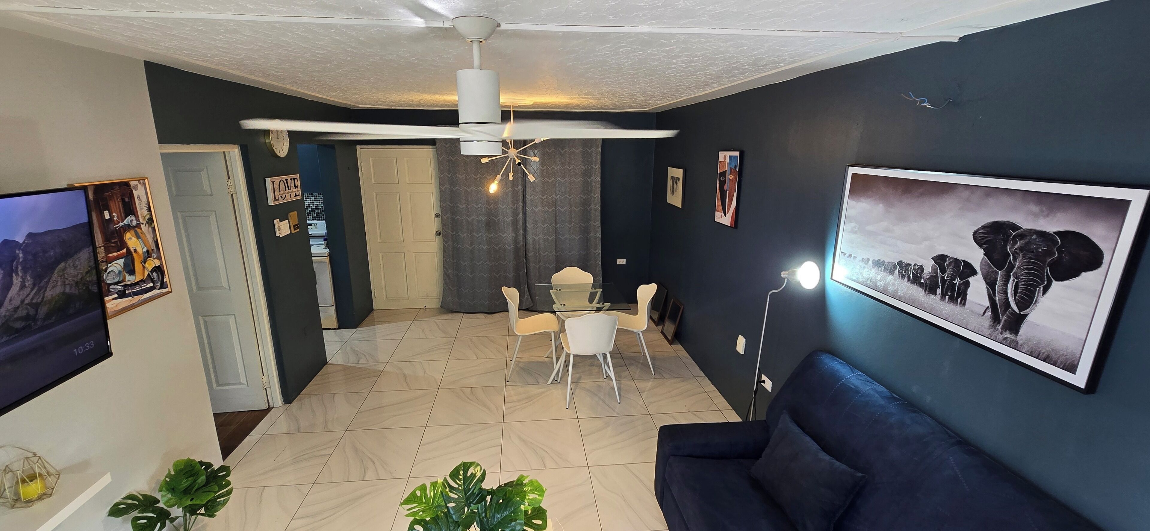A living room with a dark blue wall, a ceiling fan, a sofa, a TV, and a dining area with a table and chairs.
