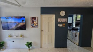 A living room with a TV, a door, a wall clock, and a kitchen visible through an open door. - The Central Comfort in Kingston (Kingston)
