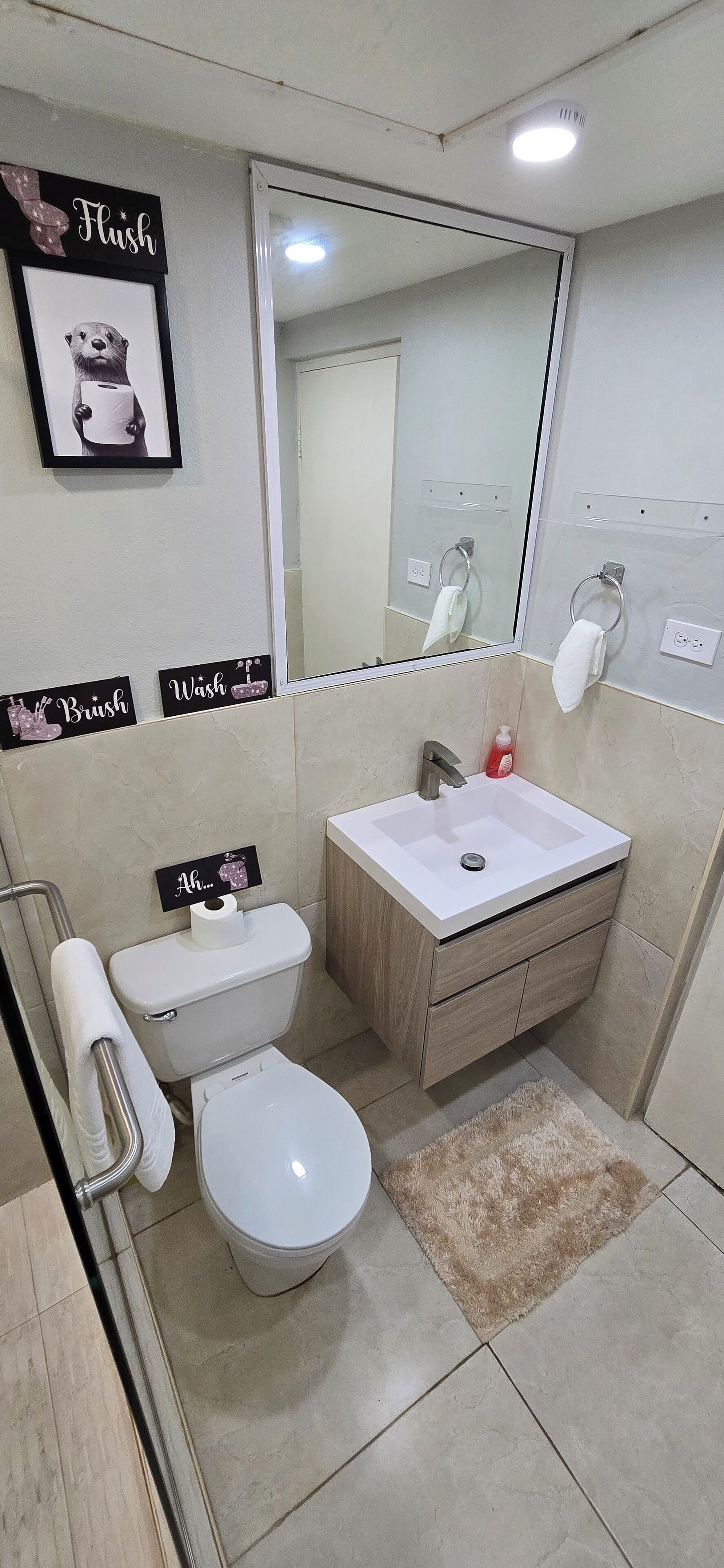 A compact bathroom with a toilet, sink, and a mirror. There are signs on the wall, a towel rack, and a small rug on the floor.