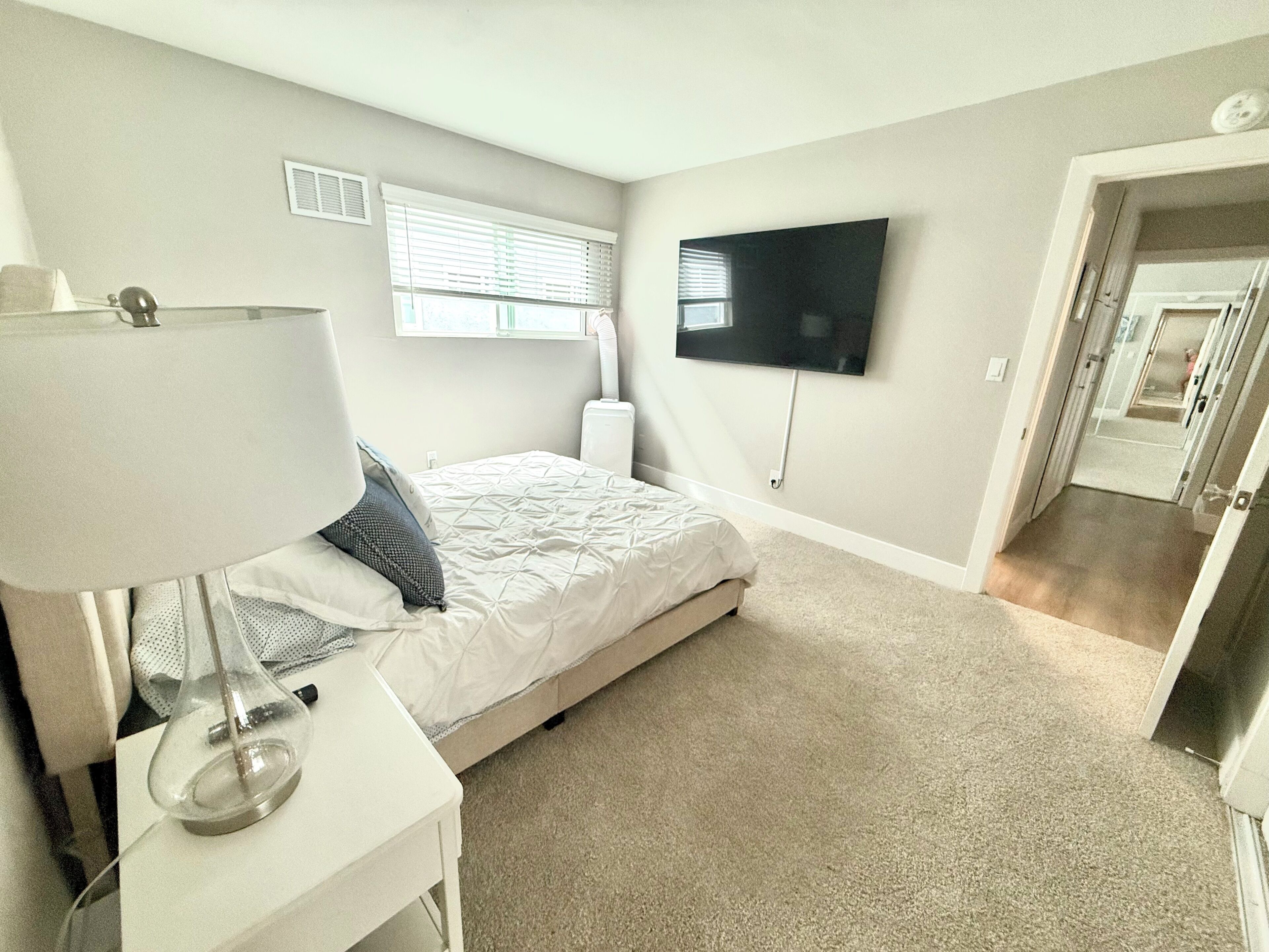 A bedroom with a bed, a TV mounted on the wall, a lamp on a bedside table, and a door leading to another room.