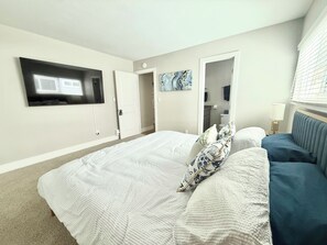 Une chambre à coucher comprenant un lit, un téléviseur fixé au mur, une porte donnant sur une salle de bain et une fenêtre avec des stores.