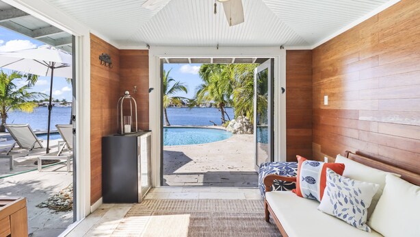 Une chambre avec vue sur une piscine et une plage, des murs en bois, un ventilateur de plafond et un canapé agrémenté de coussins à motifs.