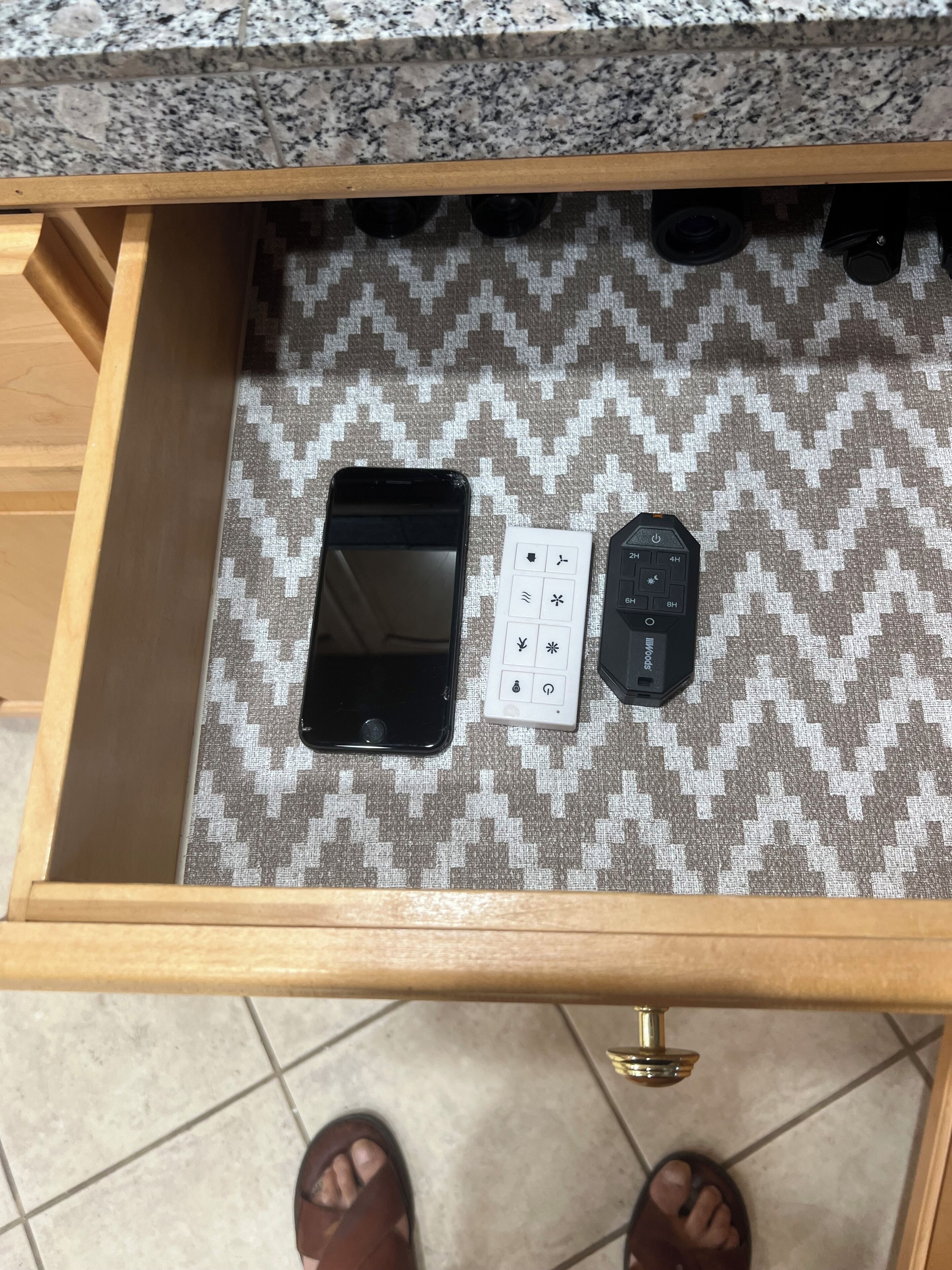 A drawer with a patterned fabric interior, containing a smartphone, a remote control, and a small white device with buttons.