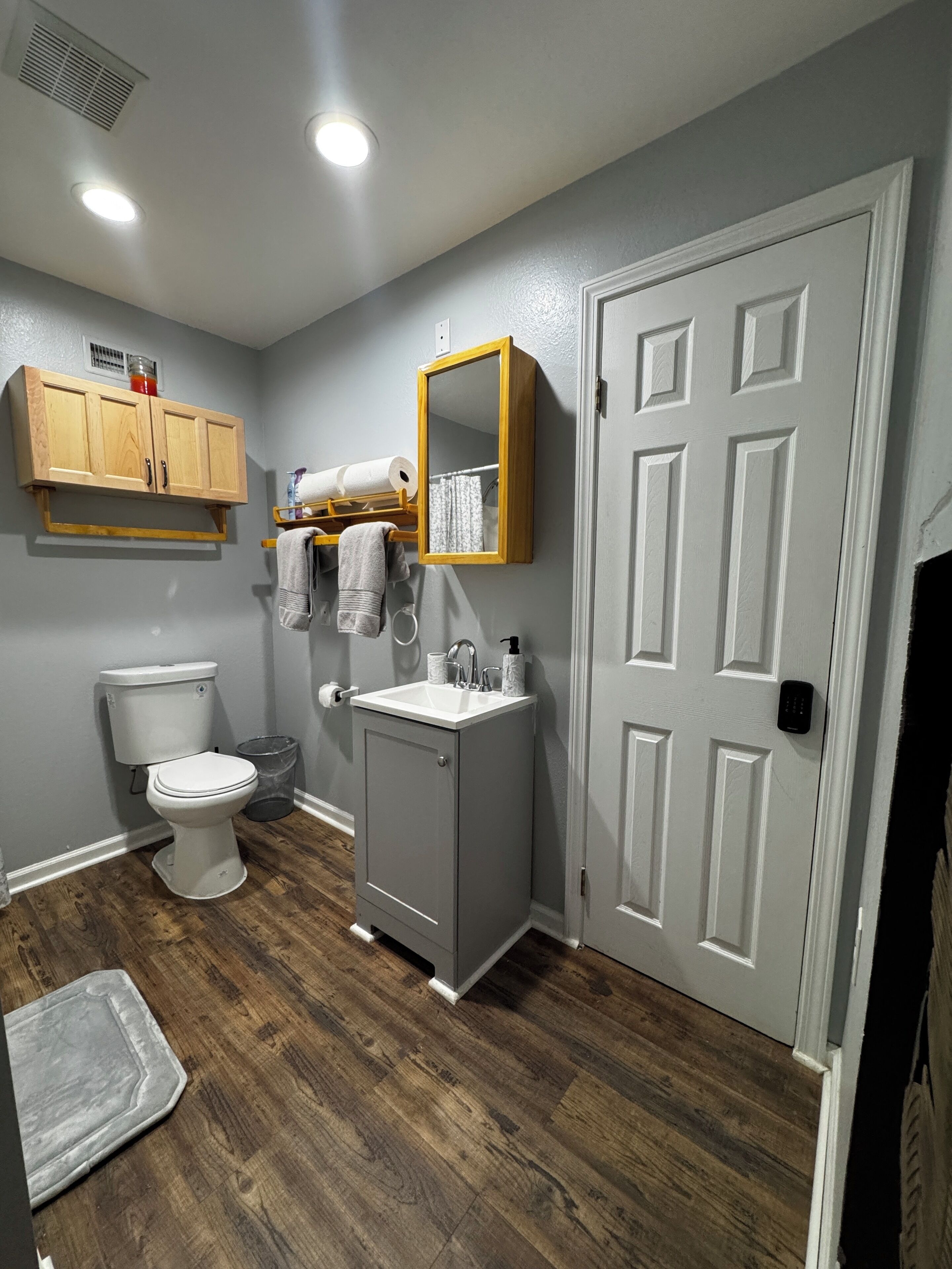 A bathroom with a toilet, sink, cabinet, mirror, and wooden flooring.