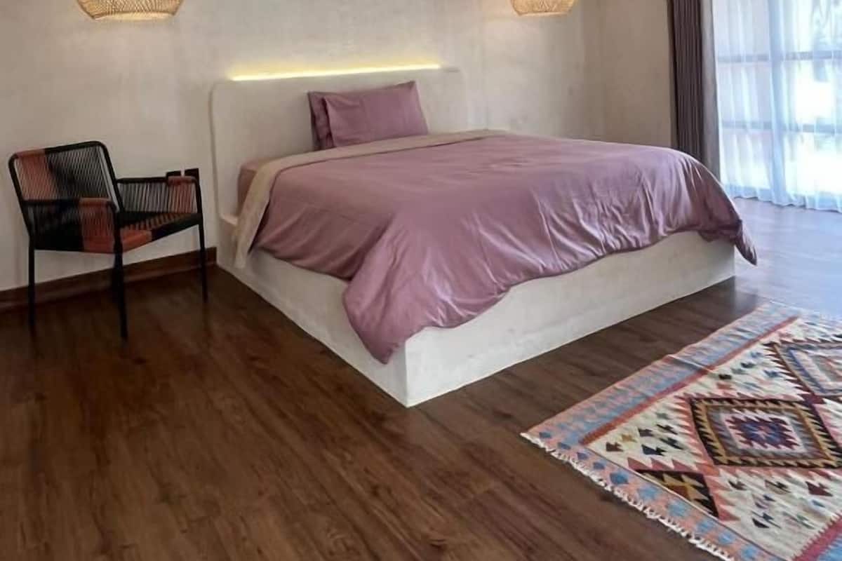 Ein modernes Schlafzimmer mit einem großen Bett, zwei Pendelleuchten, einem dekorativen Schädel, einem Sessel und einem gemusterten Teppich.