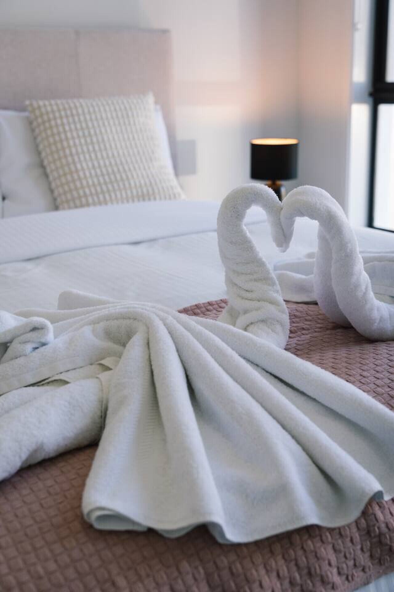 A neatly made bed with white linens and a heart-shaped towel arrangement on a pink bedspread.