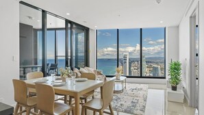A modern dining area with a large table and chairs, a view of the city and sea through large windows, and a potted plant on a windowsill.