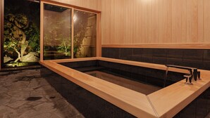 Una stanza spa con una vasca da bagno in legno, un pavimento in pietra e vista su un giardino roccioso attraverso una porta scorrevole.