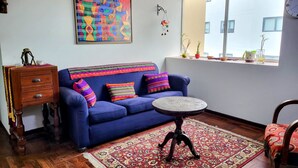 A living room with a blue sofa, a wooden side table, a round coffee table, a patterned rug, and a large abstract painting on the wall. - Apartment 3 Bedrooms | next to Delgado Clinic in Miraflores (Miraflores)