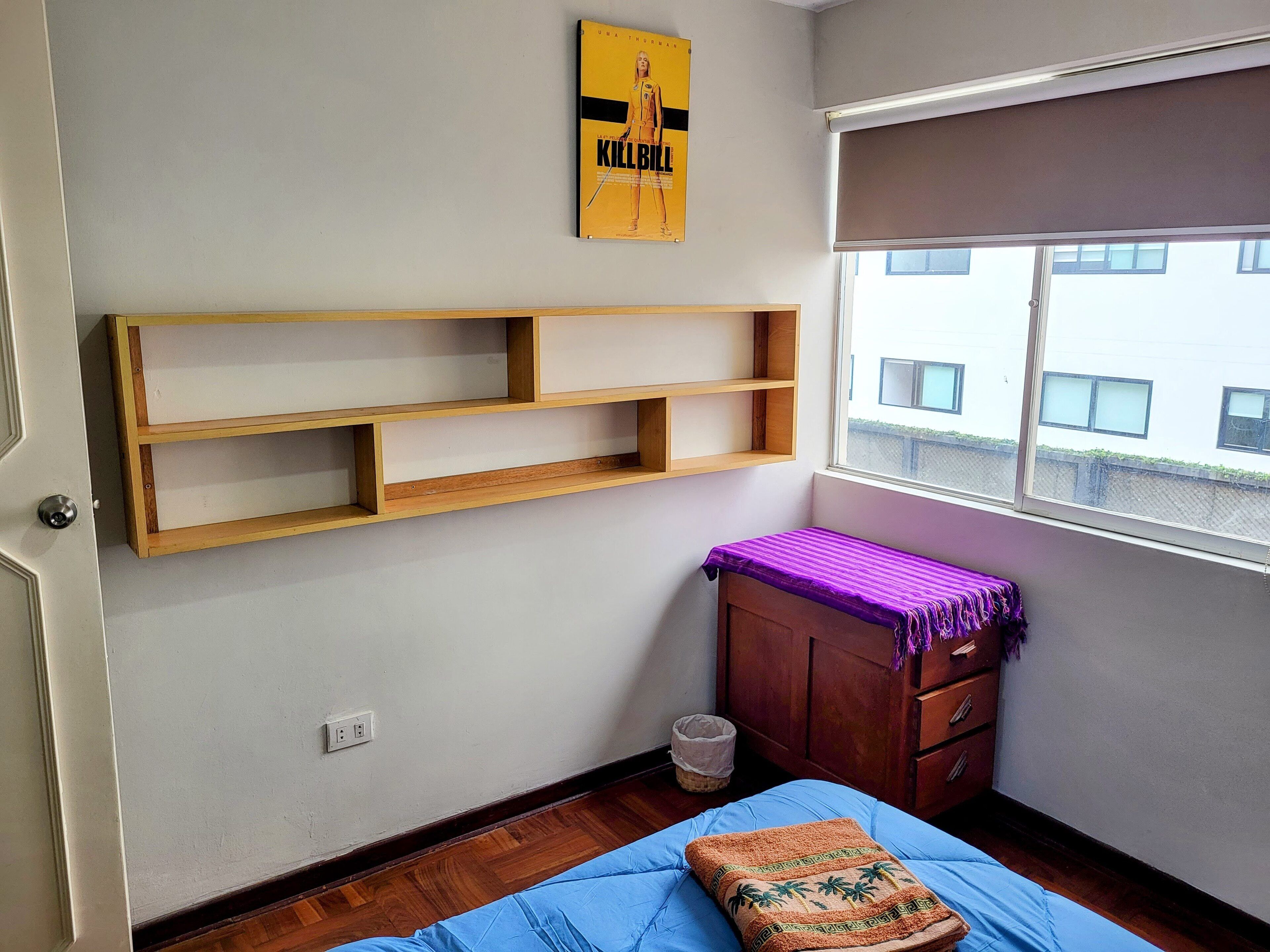 A room with a bed, a wooden shelf unit, a small cabinet, a window with a view of buildings, and a poster on the wall.