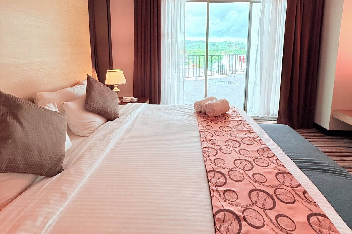 Una habitación de hotel con una cama grande, una mesita de noche con lámpara y vistas a un paisaje verde por la ventana.
