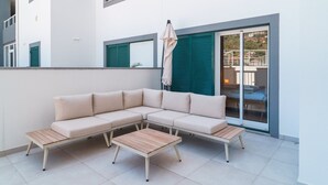 Una moderna area salotto esterna con un divano e due tavoli di legno, un ombrellone grande e una porta a vetri scorrevoli che dà su un balcone.