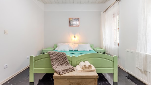 A room with a green bed, a bedside table with towels, a wall-mounted light, and a window with sheer curtains. - Nora Guest House 2BR & Garden (Nagygörbő)