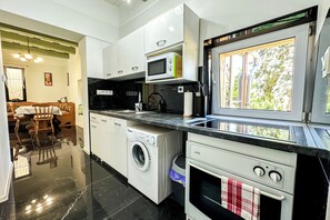 A modern kitchen with a washing machine, microwave, oven, and a window with a view of greenery. - Nora Guest House (Nagygörbő)
