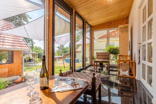 A modern interior with large windows, a wooden ceiling, and a dining area with a table, chairs, and a bottle of wine. - Nora Guest House 2BR & Garden (Nagygörbő)