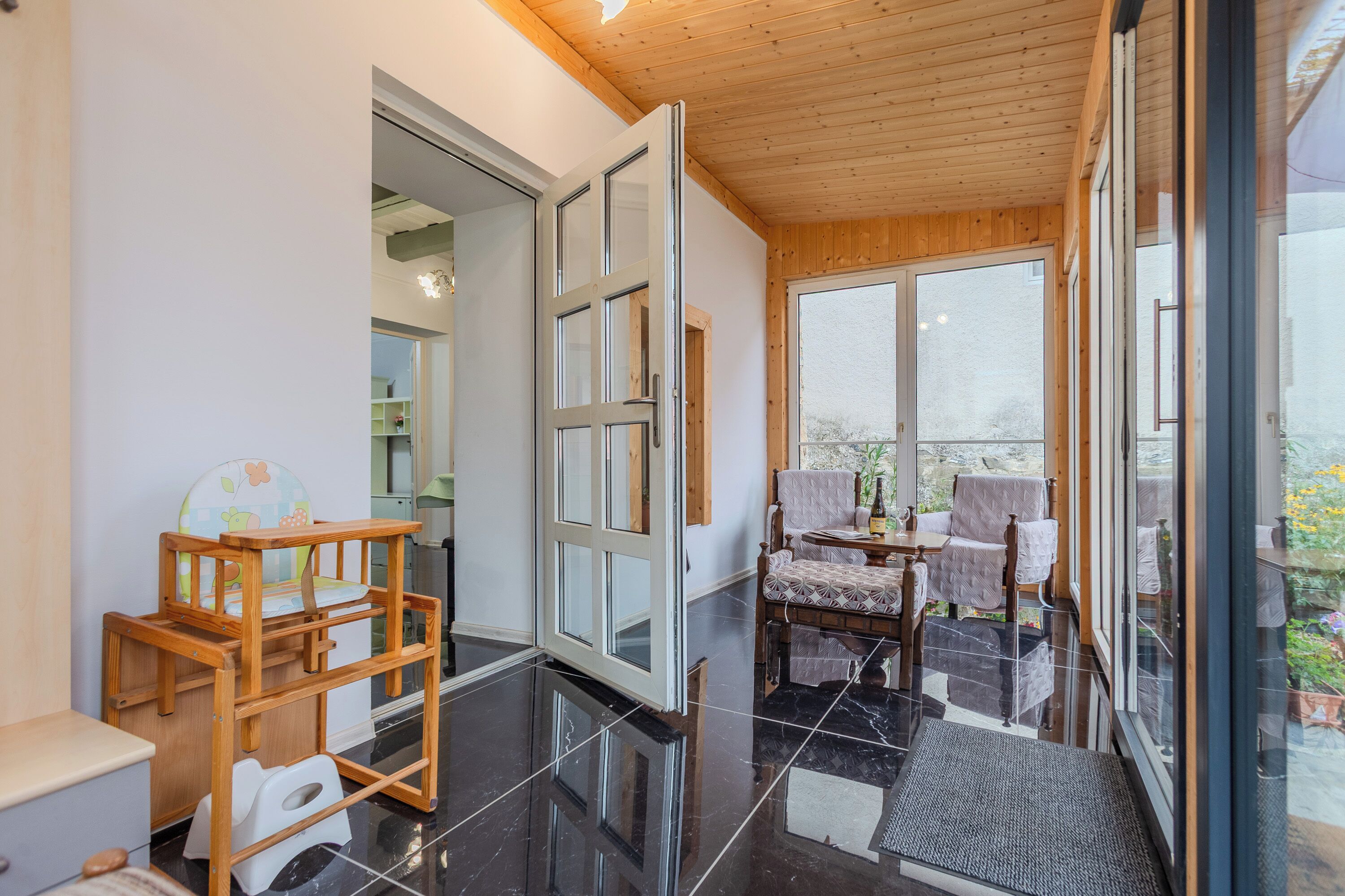 A modern interior with a wooden ceiling, a glass door leading outside, a dining area with a table and chairs, and a child's play area with a chair and a toy.