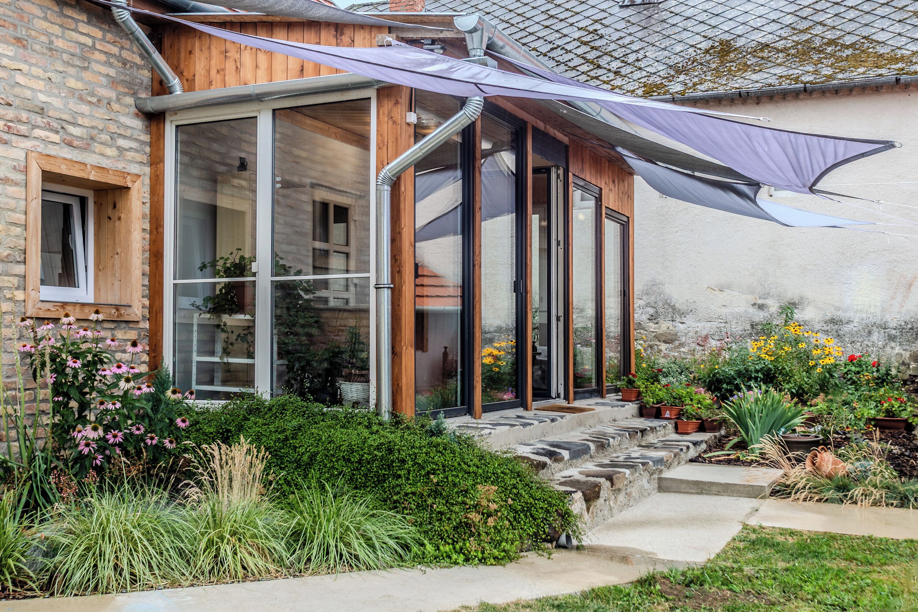 A house with a glass conservatory, wooden exterior, and a garden with various plants and flowers.