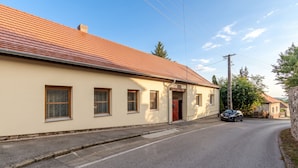 A residential street with a single-story house featuring a red-tiled roof, a red door, and a small driveway. - Nora Guest House 2BR & Garden (Nagygörbő)