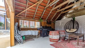 A rustic interior with a wooden ceiling, a table with a tablecloth, a hanging chair, and a grill area. - Nora Guest House 2BR & Garden (Nagygörbő)