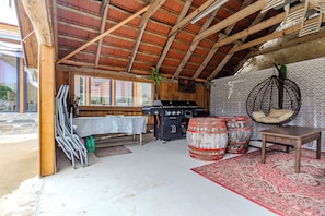 Ein rustikales Interieur mit Holzbalkendecke, einem Tisch mit Tischdecke, einem Hängekorbstuhl und einem Grillbereich.