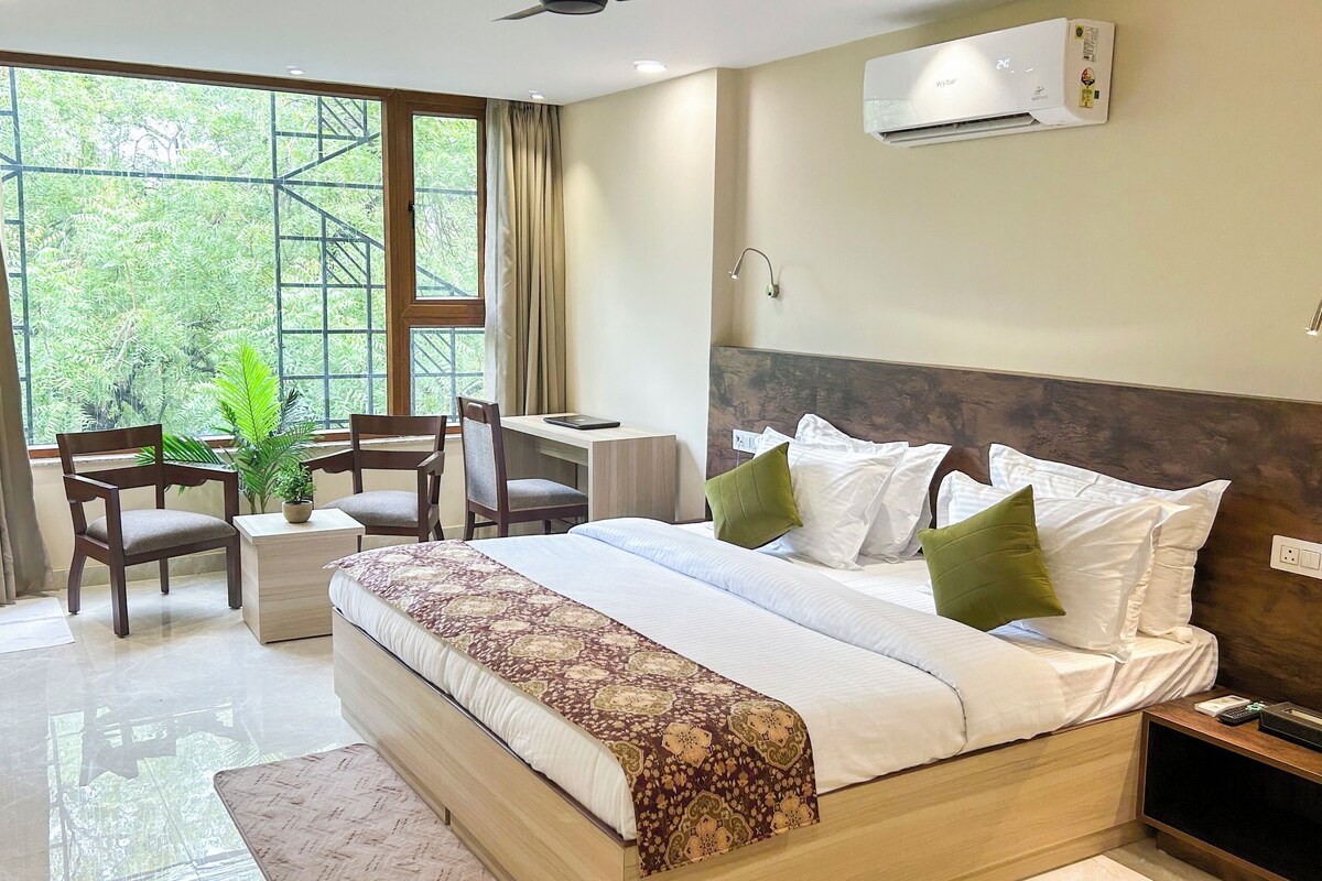 A hotel room with a bed, a desk, two chairs, a potted plant, and a window with curtains.
