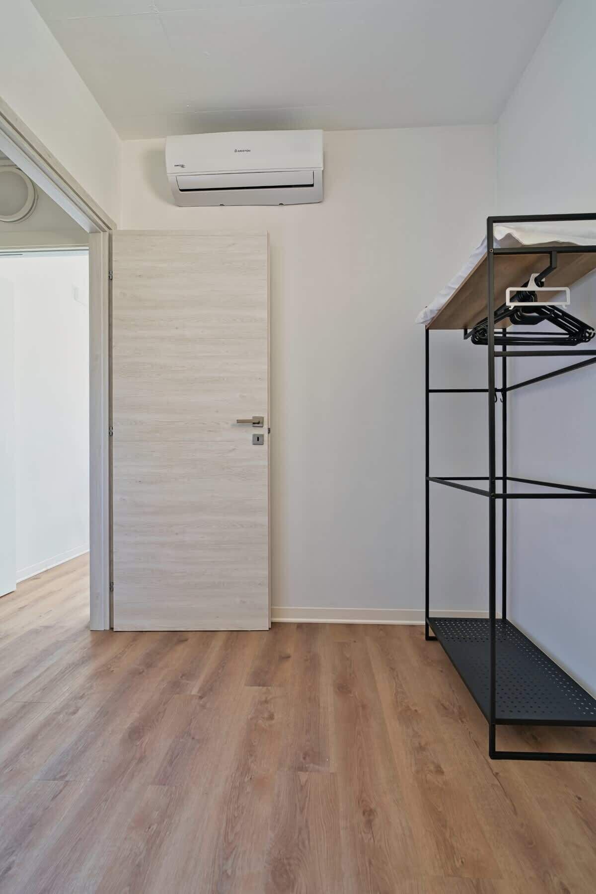 Een kamer met een houten deur, een zwart metalen rek en een airconditioner aan de muur.