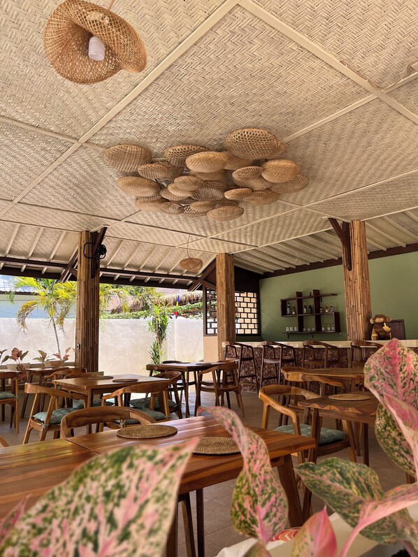 A restaurant with wooden tables and chairs, a woven ceiling, and a view of greenery outside. - Gila House (Gili Air)