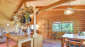 Ein Holzblockhaus-Interieur mit einem Holzesstisch, Stühlen, einer Küchenzeile mit hängenden Töpfen, einem großen Fenster und einem farbenfrohen Teppich.