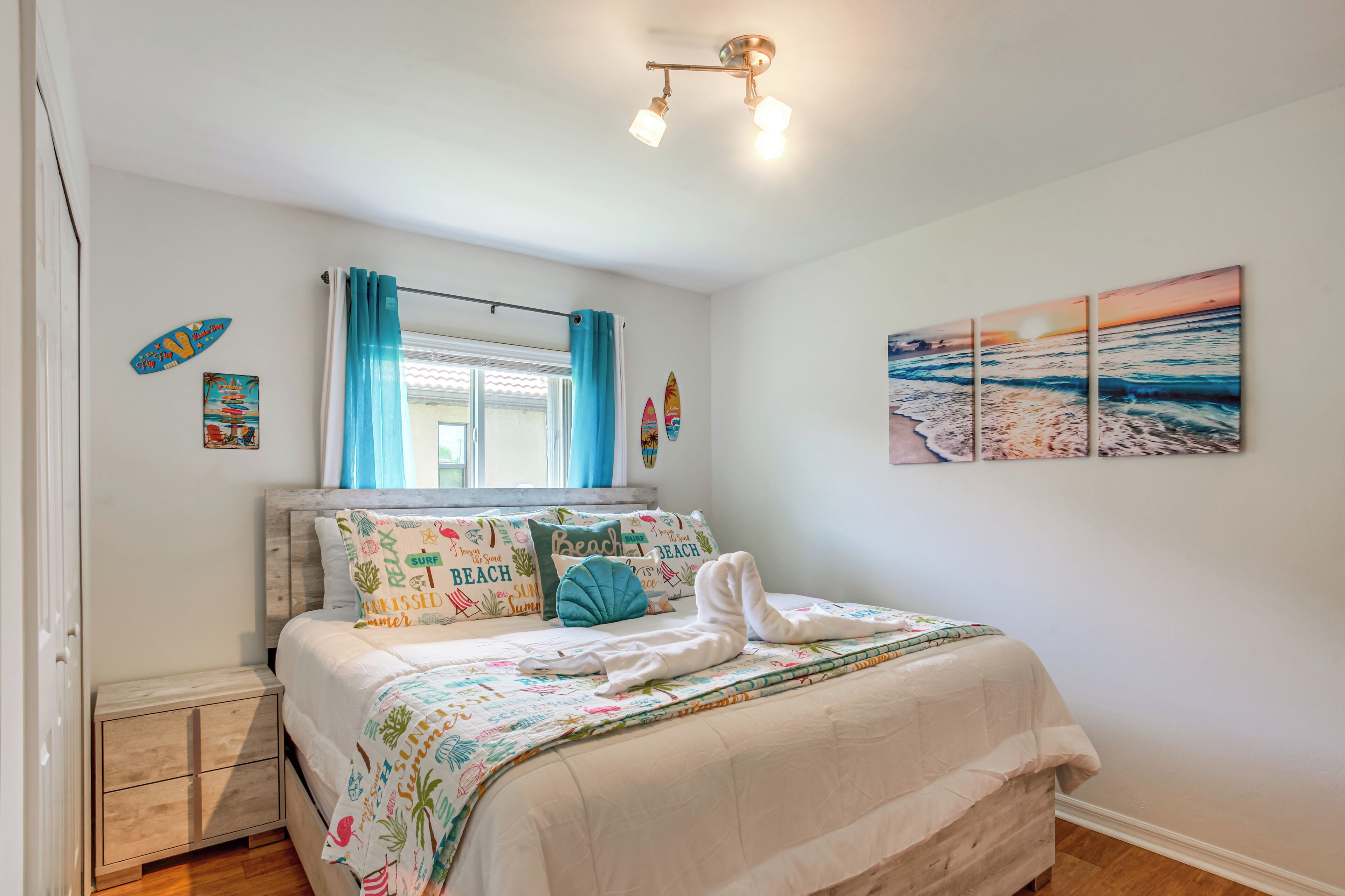 A bedroom with a bed, a nightstand, a window with curtains, a wall with a beach painting, and a ceiling light fixture.
