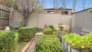 A backyard with a wooden fence, a small garden area with various plants, a yellow chair, and a table.