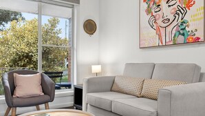 A living room with a grey sofa, a round coffee table, and a chair. There is a large window with a view of trees and a bench outside. A colorful painting is on the wall.
