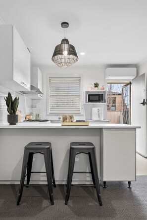 A modern kitchen with white cabinetry, a central island with stools, a hanging light fixture, and a window with blinds.
