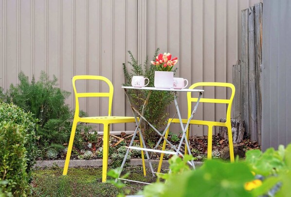 A small outdoor seating area with two yellow chairs and a table, surrounded by plants and a corrugated metal wall.