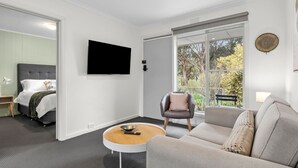 A modern living room with a grey sofa, a round coffee table, a TV mounted on the wall, and a large window with a view of greenery.