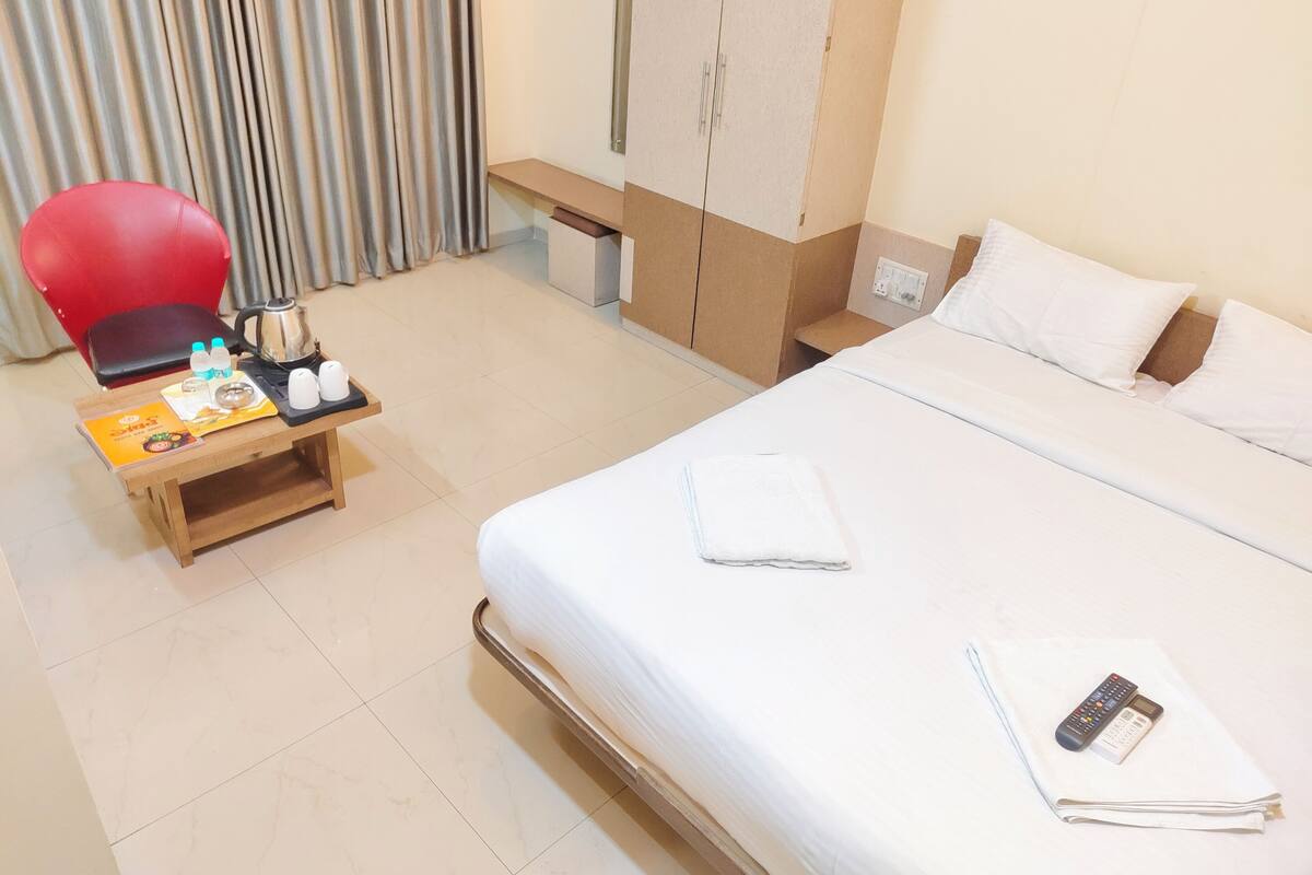 A hotel room with a bed, a desk with a chair, a red chair, a minibar, and a window with curtains.