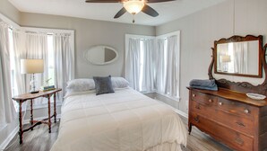 Quarto com cama, penteadeira, espelho, ventoinha no teto e janela com cortinas.