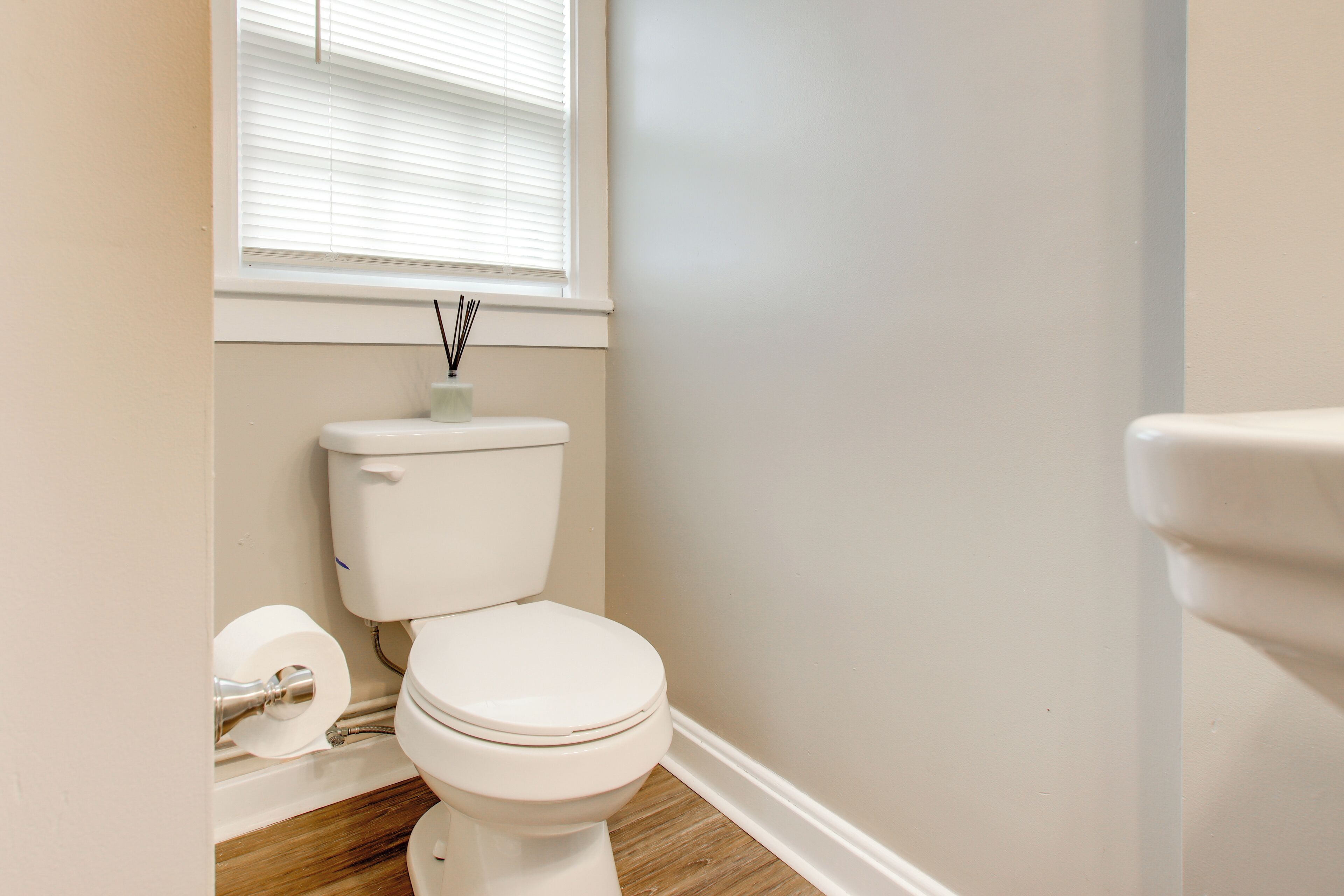 A bathroom with a toilet, wooden flooring, a window with blinds, and a wall-mounted toilet paper holder.