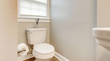 A bathroom with a toilet, wooden flooring, a window with blinds, and a wall-mounted toilet paper holder.