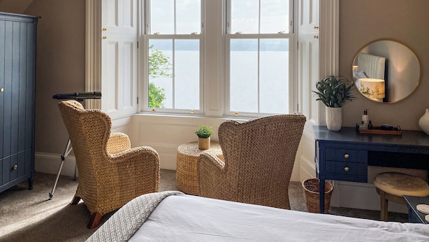 A bedroom with a large bed, two wicker chairs, a desk, a dresser, a chandelier, and a window with a view of the sea. - 3 bedroom accommodation in Blairmore, near Dunoon (Blairmore, near Dunoon)