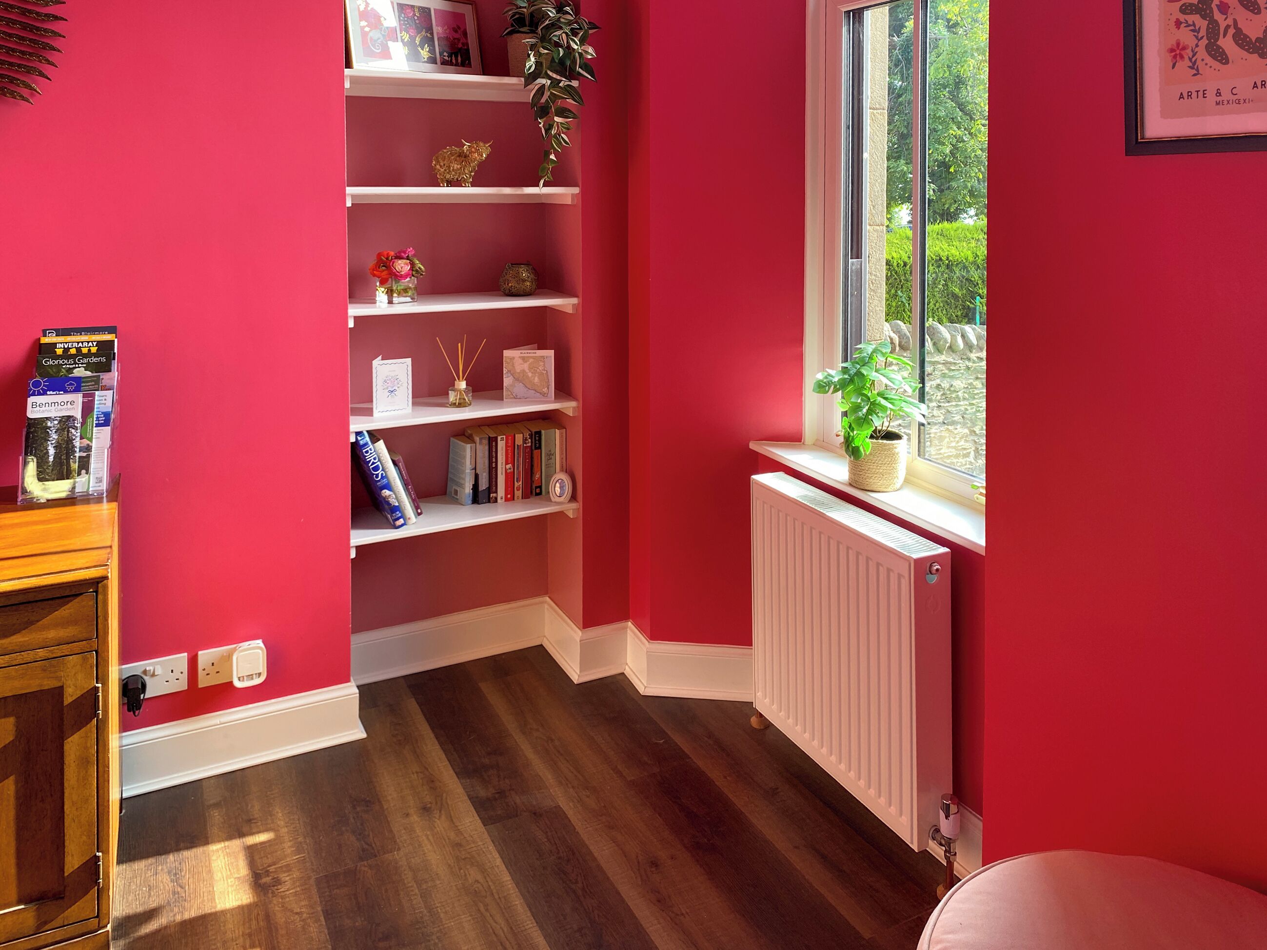 Une pièce avec un mur rouge, un radiateur blanc, une fenêtre donnant sur un paysage agréable, une bibliothèque remplie de livres et d’objets décoratifs, et un meuble en bois.