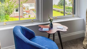 A room with a large window, a blue chair, a small wooden table with a vase of flowers, and a view of a garden and house. - 3 bedroom accommodation in Blairmore, near Dunoon (Blairmore, near Dunoon)