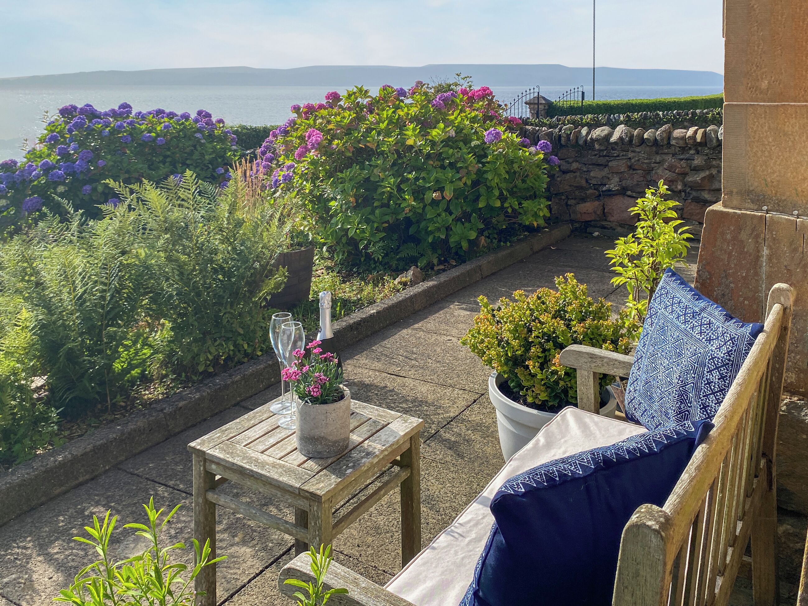 Une terrasse avec une table et des chaises en bois, une bouteille de champagne et une vue sur la mer.